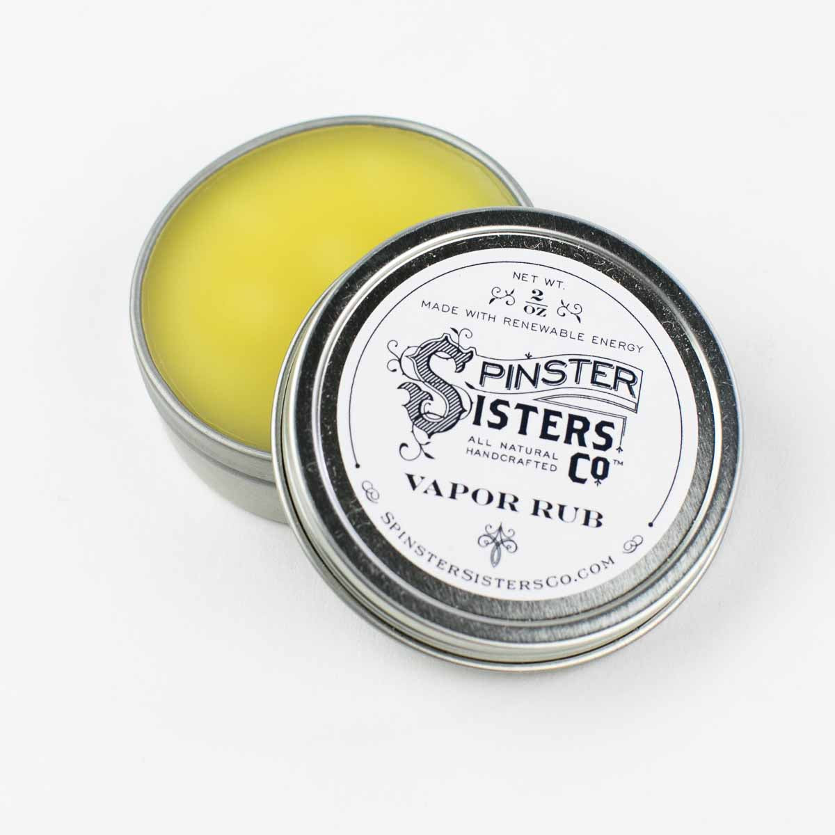 A round tin of Spinster Sisters Vapor Rub, open to show yellow balm, with the lid next to it displaying the Spinster Sisters, Co. logo and information on a white background.