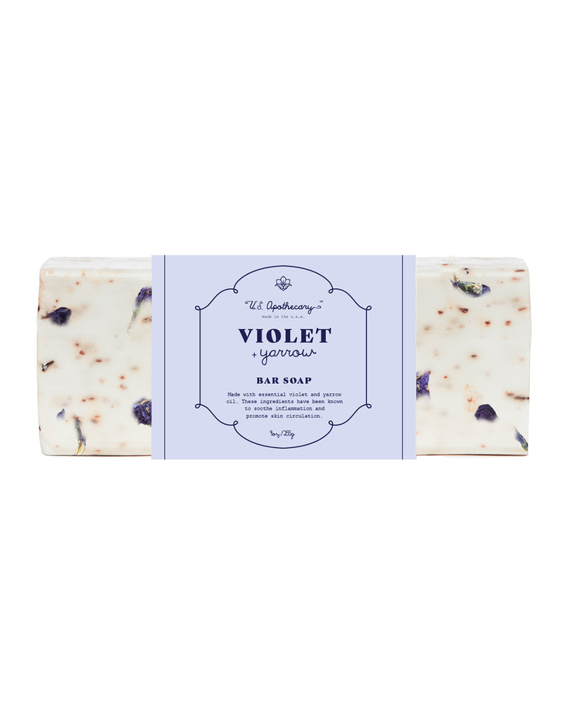 A bar of U.S. Apothecary Violet + Yarrow Triple Milled Bar Soap with visible petals and flowers embedded, labeled "violet soap" in an elegant typeface on a central blue label, enriched with shea butter.