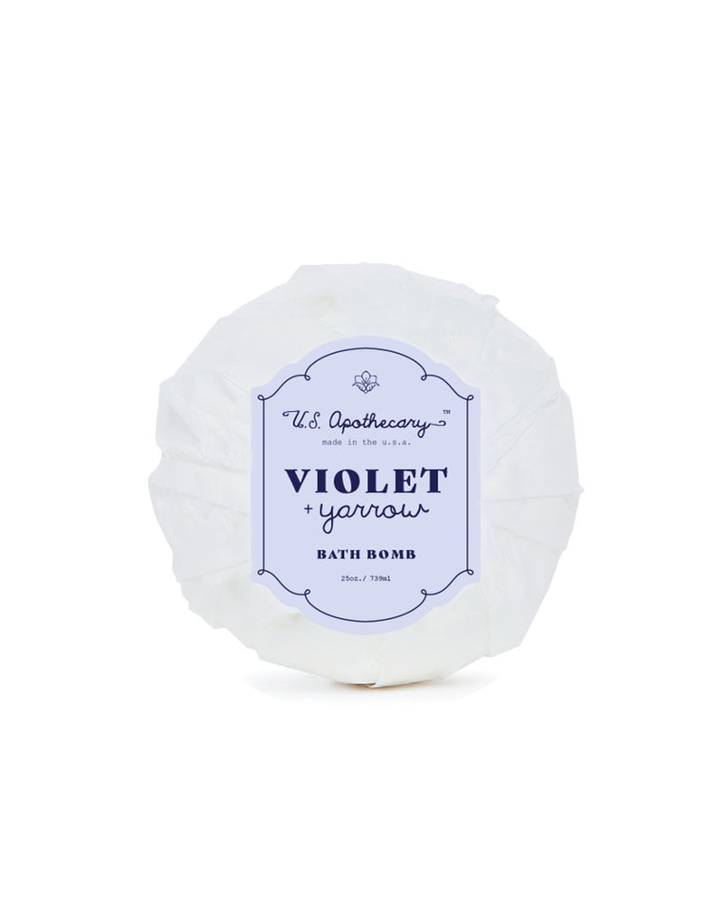 A U.S. Apothecary Violet + Yarrow Bath Bomb, designed for relaxing tense muscles, displayed in its packaging with a white and blue label, isolated on a white background.