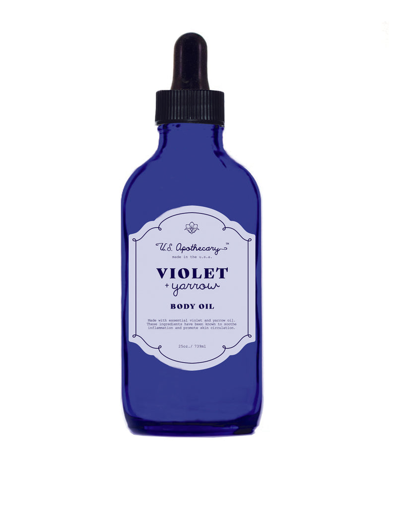 A blue glass dropper bottle labeled "U.S. Apothecary Violet + Yarrow Body Oil" from "U.S. Apothecary," set against a white background. The bottle is 2 fl oz (59ml).