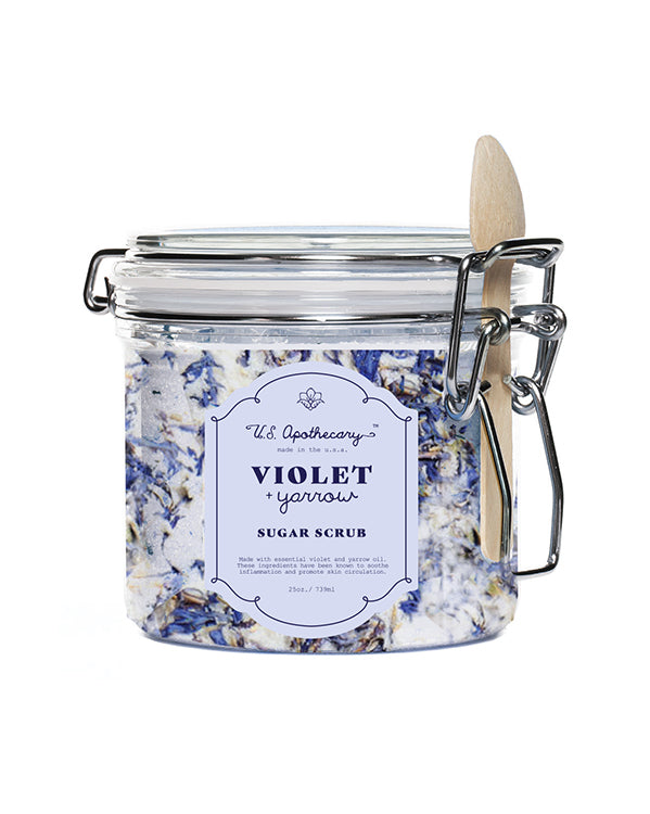 A glass jar with a metal clasp filled with U.S. Apothecary Violet + Yarrow Sugar Scrub and coconut oil, labeled "U.S. Apothecary Violet + Yarrow" by U.S. Apothecary. A wooden spoon is attached to the side.