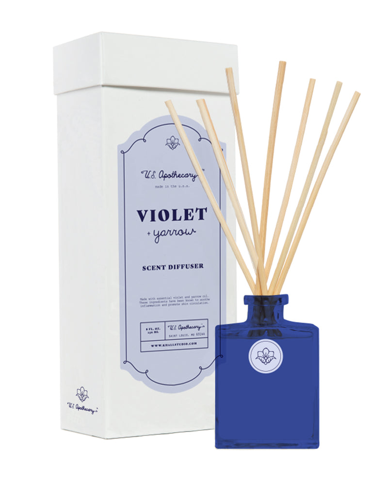 A product image featuring a U.S. Apothecary Violet + Yarrow Scent Diffuser Kit with reed sticks and a white packaging box labeled "the apothecary, violet yarrow scent diffuser.
