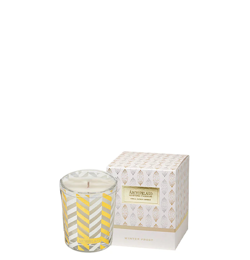 An Archipelago Botanicals Winter Frost Boxed Votive Candle in a clear glass with yellow and white diagonal stripes, next to its decorative box that has a geometric diamond pattern in white and gold, labeled "Winter Frost.