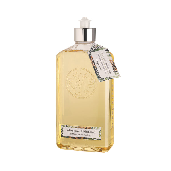 A transparent bottle of liquid soap with a label reading "Mangiacotti White Spruce Natural Kitchen Soap." The soap is yellow, and the bottle features an embossed emblem and a pump top.