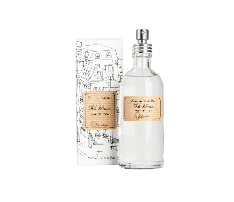 A clear glass perfume bottle with a label reading "eau de toilette, Lothantique White Tea" next to its cardboard packaging featuring a sketched cityscape design.