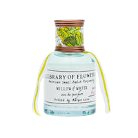 A bottle of Margot Elena's Library of Flowers Willow & Water Eau De Parfum, with a clear blue liquid, adorned with a decorative white label featuring floral artwork and a yellow ribbon tied around the.