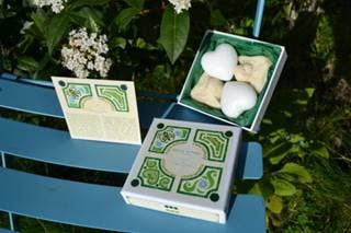 Two Senteurs De France decorative boxes on a blue bench, one open displaying three white ceramic hearts nestled in fabric. Lush greenery and white flowers in the background, evoking thoughts of French garden soaps.