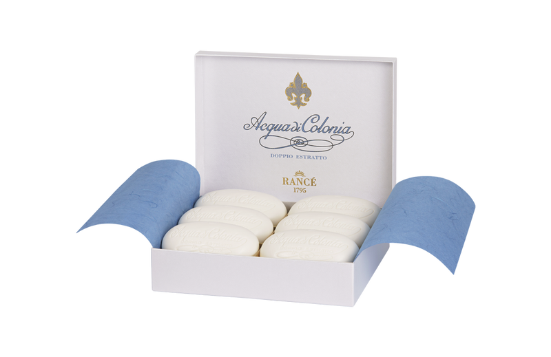 A pair of hands wearing blue gloves holds an open white box displaying oval-shaped Rancé Classic Soaps labeled "Acqua di Colonia" with a citrus scent against a white background.