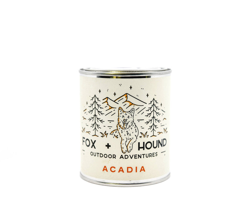 A white, cylindrical Fox + Hound Acadia Odor Eliminator Soy Candle from the National Park Series with a label featuring an illustrated forest scene and a fox. The text reads "Fox and Hound Outdoor Adventures - Acadia National Park." The background is solid white.