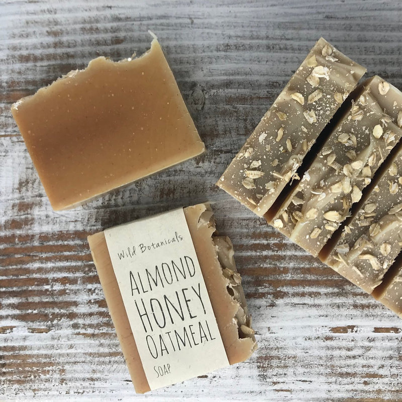 Handmade Wild Botanicals Almond Honey Soap bars laid on a wooden surface, some with visible oat flakes on top. One bar has a wildflower seed paper label.