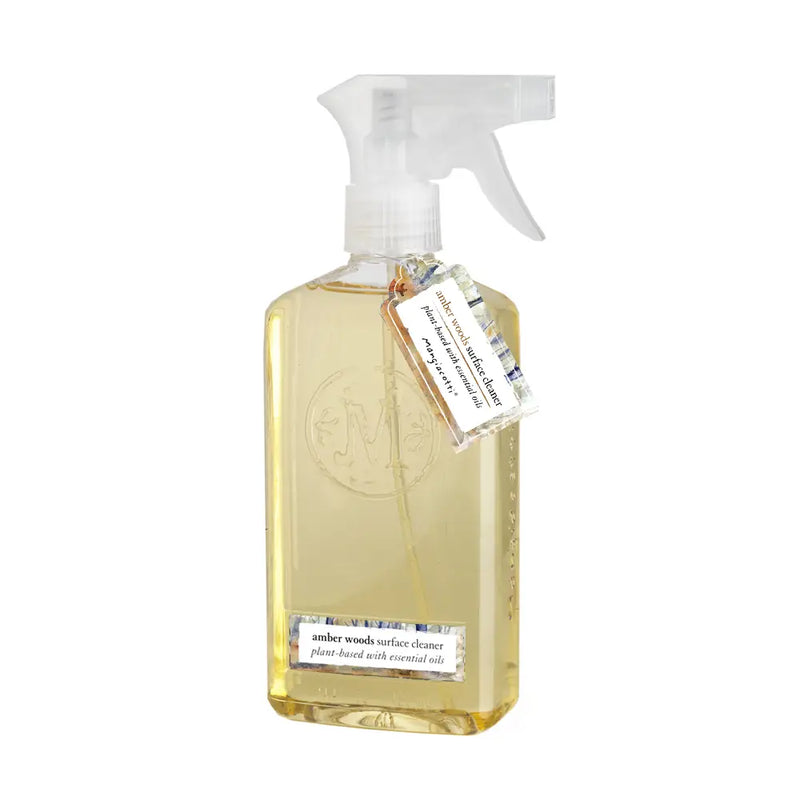 A transparent spray bottle with Mangiacotti Amber Woods Surface Cleaner, featuring an embossed Mangiacotti logo and a label tied with a string around the neck, suitable for non-porous surfaces.