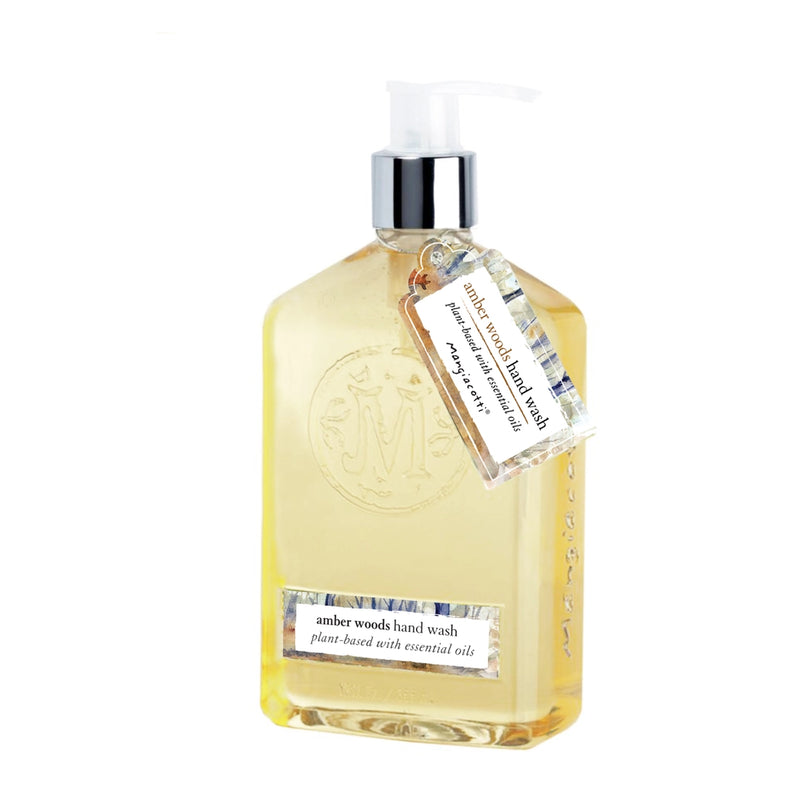 A clear bottle of Mangiacotti Amber Woods Hand Wash with a pump dispenser, labeled "amber woods hand wash." The bottle features an elegant monogram and a tag describing the product as plant-based hand wash with essential oils.