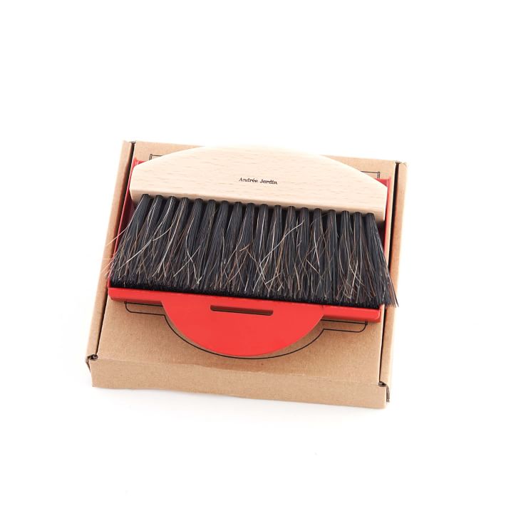 A Andrée Jardin Mr. & Mrs. Clynk Natural Table Brush & Dustpan Set - Red with a wooden handle, resting in a cardboard box with a red base. The brush, made from 100% natural fibers, appears suitable for grooming or cleaning tasks.