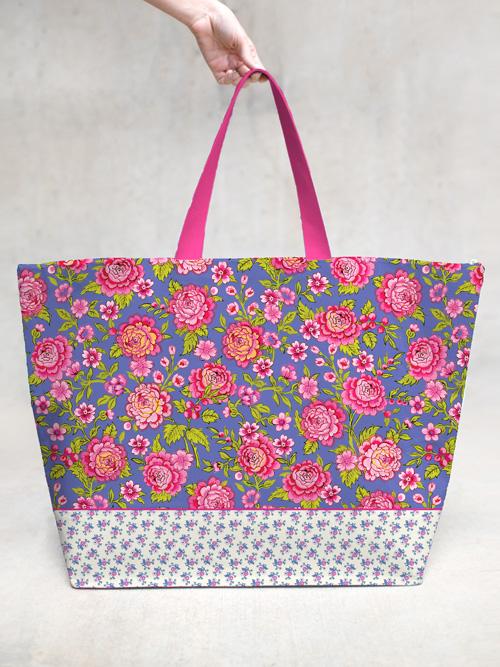 A hand holding a Margot Elena TokyoMilk Anthemoessa Large Tote Bag with a vibrant floral pattern in shades of pink, red, and blue against a gray background. The bag features contrasting woven handles and a decorative white and blue border.