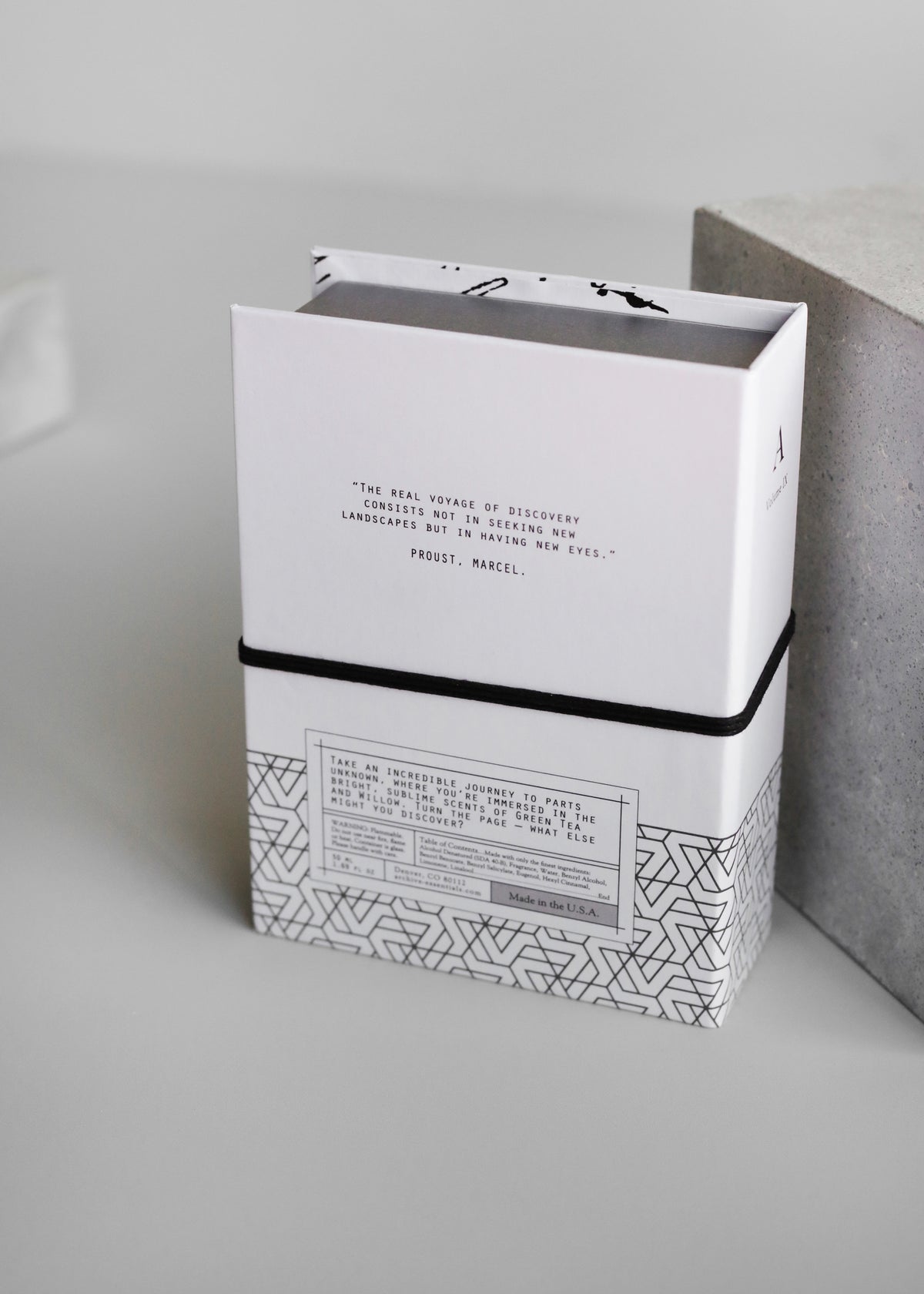 A stack of white books with black and grey geometric patterns on a light grey surface. The visible book cover quotes Marcel Proust and features a willow design from the ARCHIVE by Margot Elena - Journey Within Fragrance collection by Margot Elena.