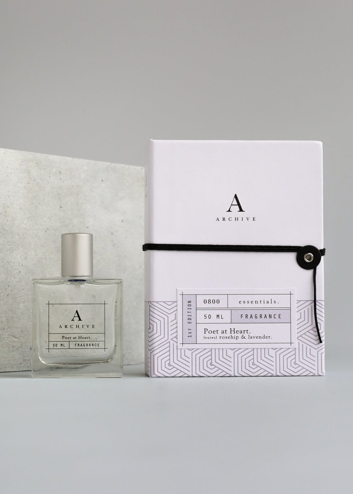 A square Poet at Heart Fragrance bottle with a label next to a white box adorned with geometric patterns and placed on a gray tabletop. The box has an "a" logo and text describing the calming scent by Margot Elena.
