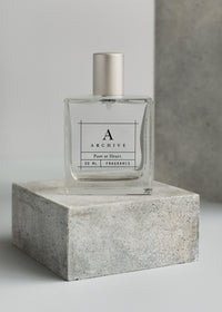A clear glass perfume bottle labeled "ARCHIVE by Margot Elena - Poet at Heart Fragrance 50 ml lavender rosehip" resting on a grey concrete pedestal against a light grey background.