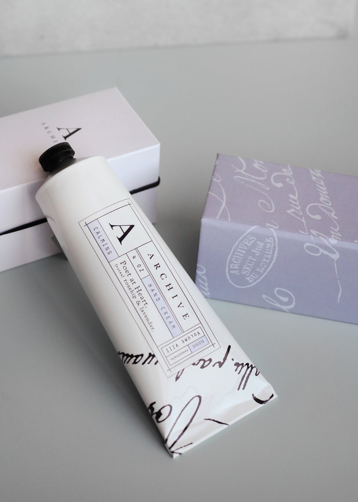 An elegant nourishing Poet at Heart Hand Cream tube by Margot Elena on a gray surface next to two neatly stacked light-colored boxes with cursive text. The tube displays a minimalist black and white design with the letter "a" prominent.