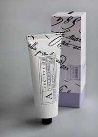An elegant skincare tube of ARCHIVE by Margot Elena - Poet at Heart Hand Cream packaged in a similarly designed box with sophisticated black script on a white background. The tube and box are set against a soft grey backdrop.