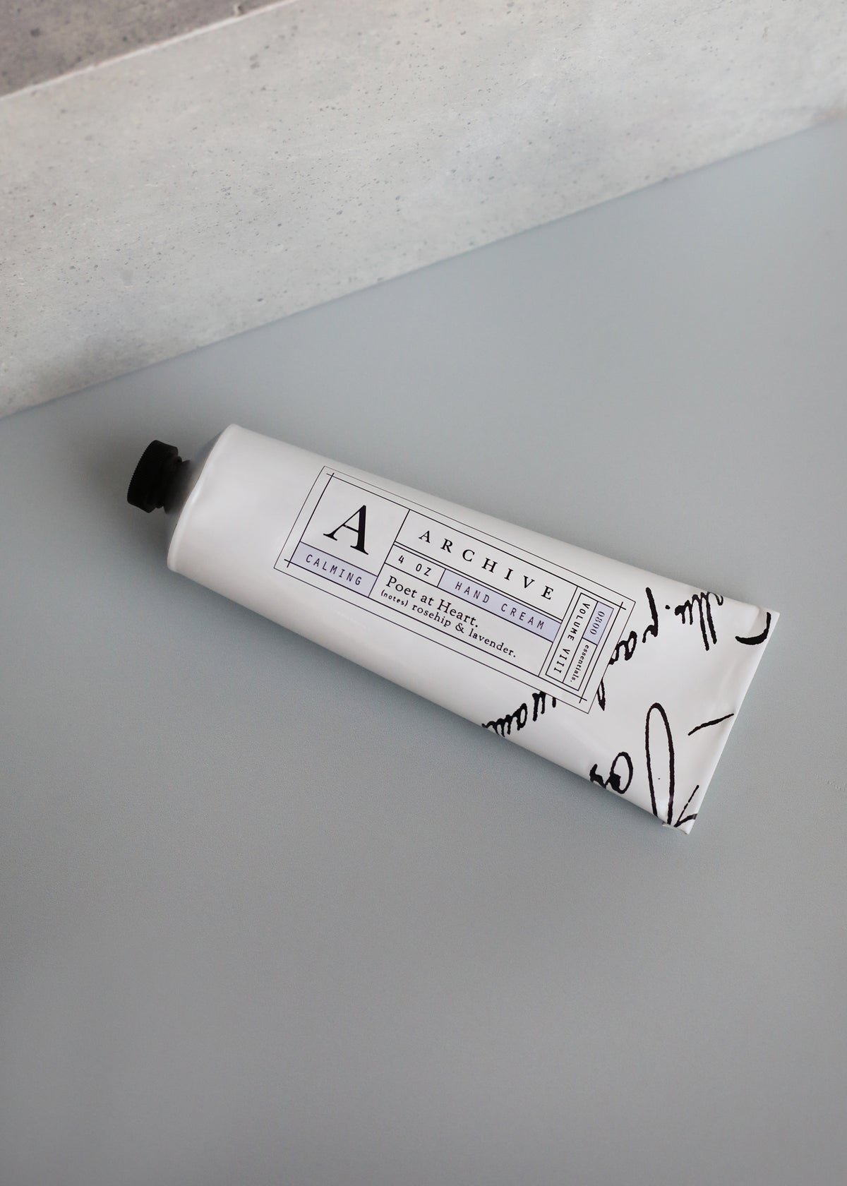A tube of ARCHIVE by Margot Elena - Poet at Heart Hand Cream with a sleek, artistic design, labeled "archive," featuring nourishing hand cream and resting on a light gray surface with a subtle concrete texture.