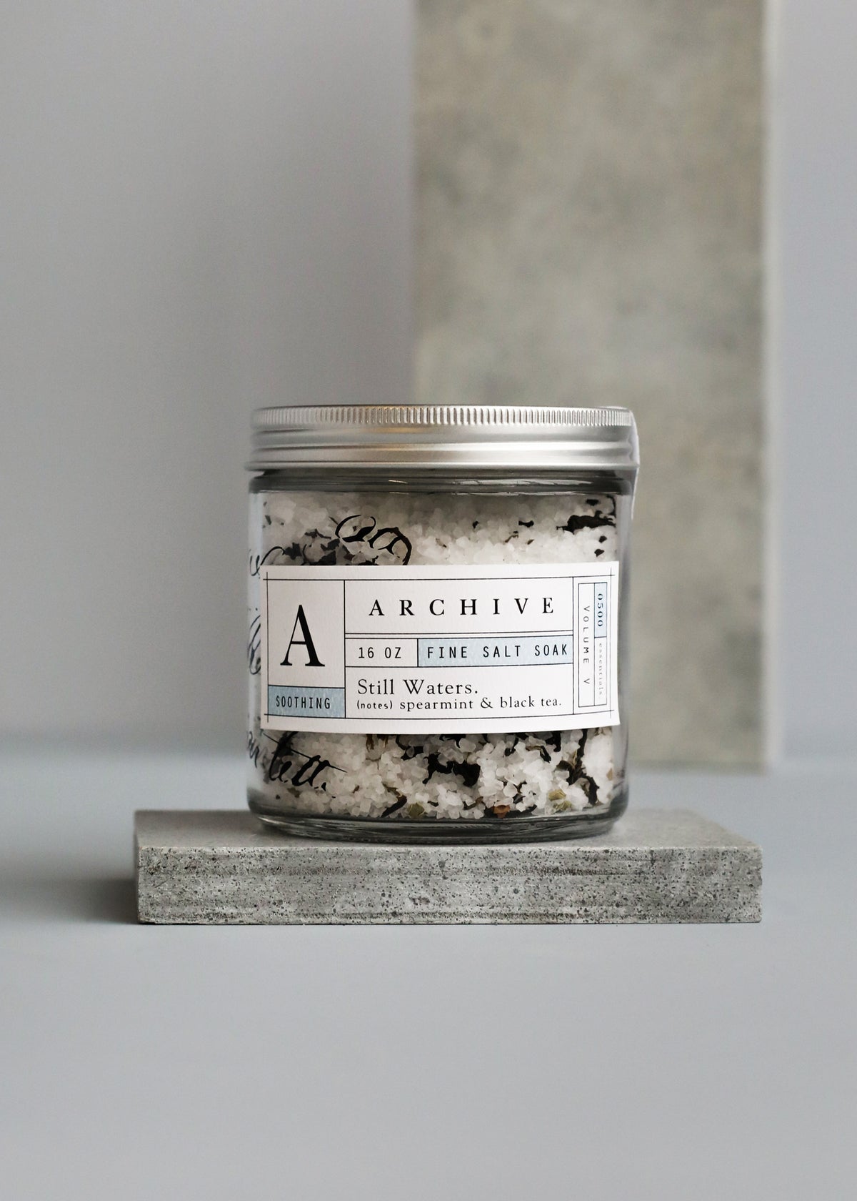 A jar of Margot Elena's ARCHIVE Still Waters Salt Soak, with spearmint and black tea, displayed on a gray stone platform against a neutral backdrop.