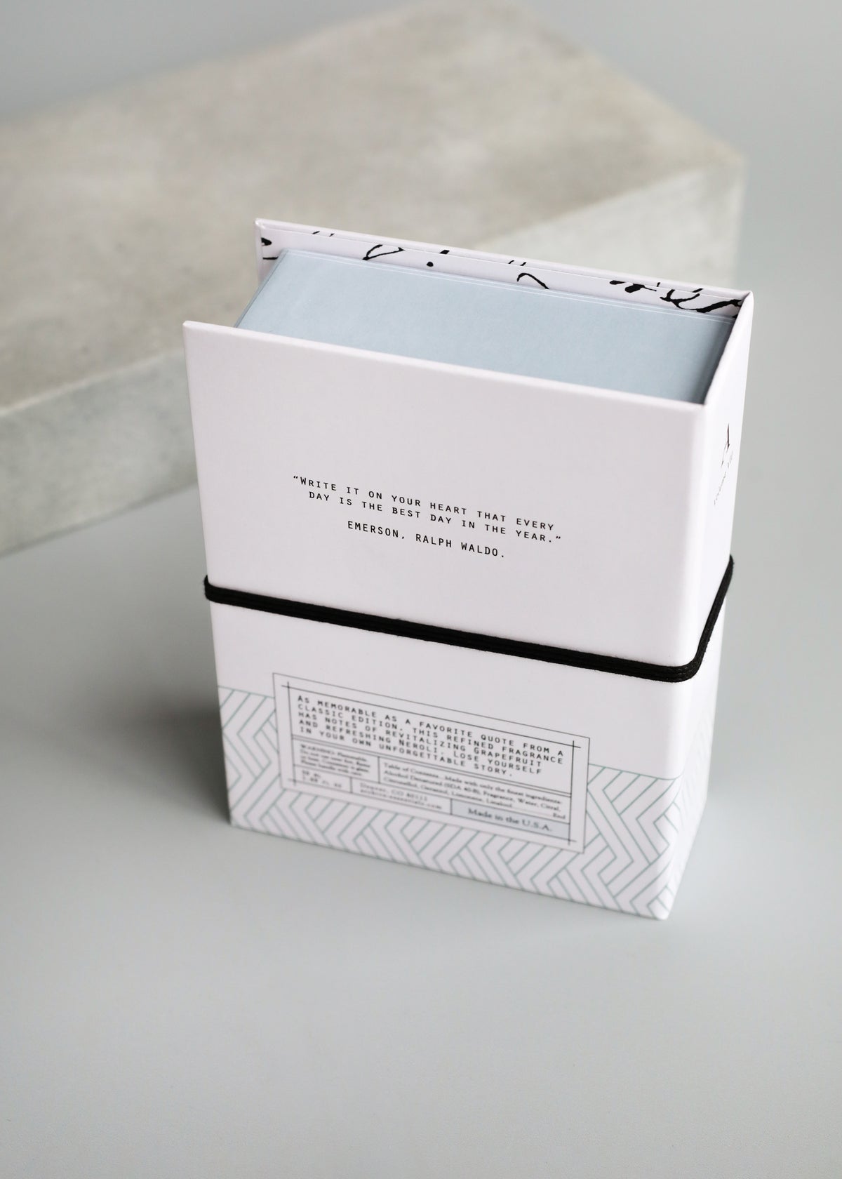 Two stacked books with hard covers. The top book is open, displaying a quote by Ralph Waldo Emerson. The bottom book, encased in a keepsake box, features a geometric design on its ARCHIVE by Margot Elena - Yet to be Written Fragrance.
