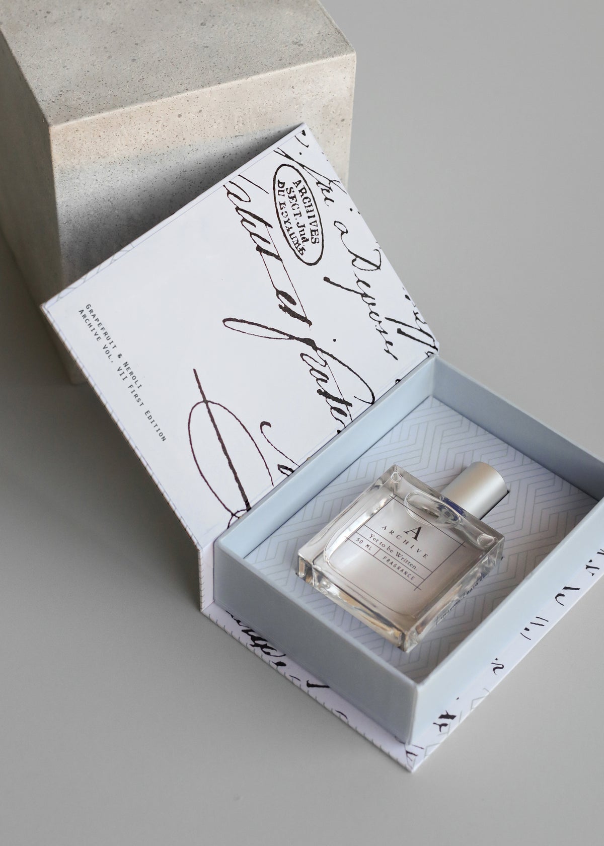 A glass perfume bottle sits inside an open light blue keepsake box with a matching outer box nearby, all featuring elegant, cursive writing and a geometric pattern inside the box. The product is ARCHIVE by Margot Elena - Yet to be Written Fragrance.