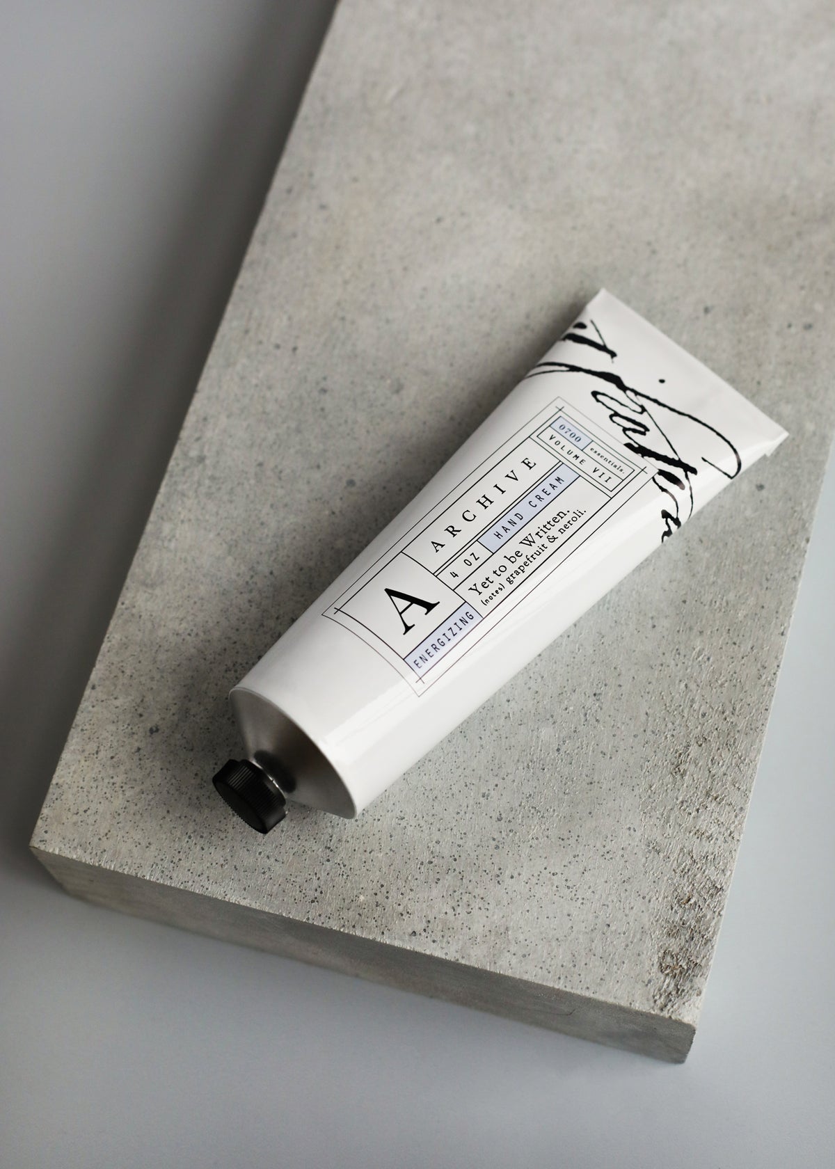 A minimalist tube of ARCHIVE by Margot Elena - Yet to be Written Hand Cream with elegant black and white labeling, featuring revitalizing citrus, resting on a gray stone surface with a soft shadow.