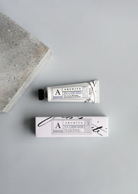 Top view of a Yet to be Written Hand Cream tube from ARCHIVE by Margot Elena next to its packaging box, both featuring elegant black and white designs with revitalizing citrus accents, set on a plain grey background with a concrete block to one side.