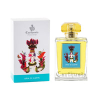 A bottle of Carthusia Aria di Capri Eau de Parfum by Carthusia I Profumi de Capri next to its box. The bottle has a clear square shape with a yellow liquid inside and a gold cap. The box is white with a colorful floral and emblem.