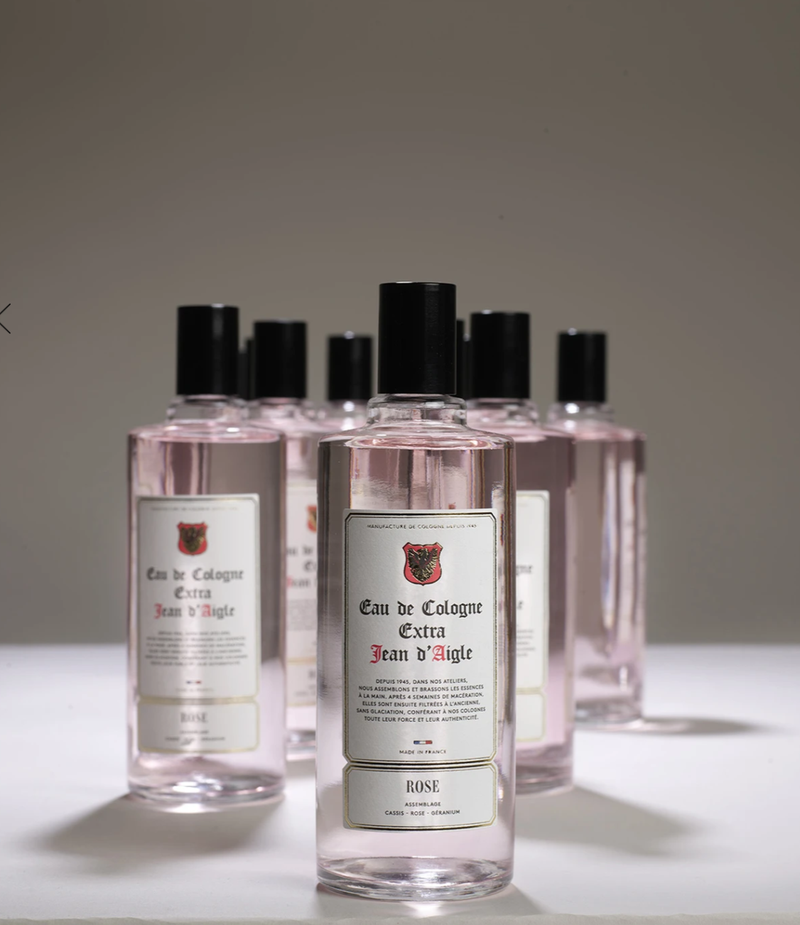 Five bottles of Jean d'Aigle Rose Eau De Cologne showcased with a focus on the front bottle and blurred ones in the background, on a neutral gray backdrop.