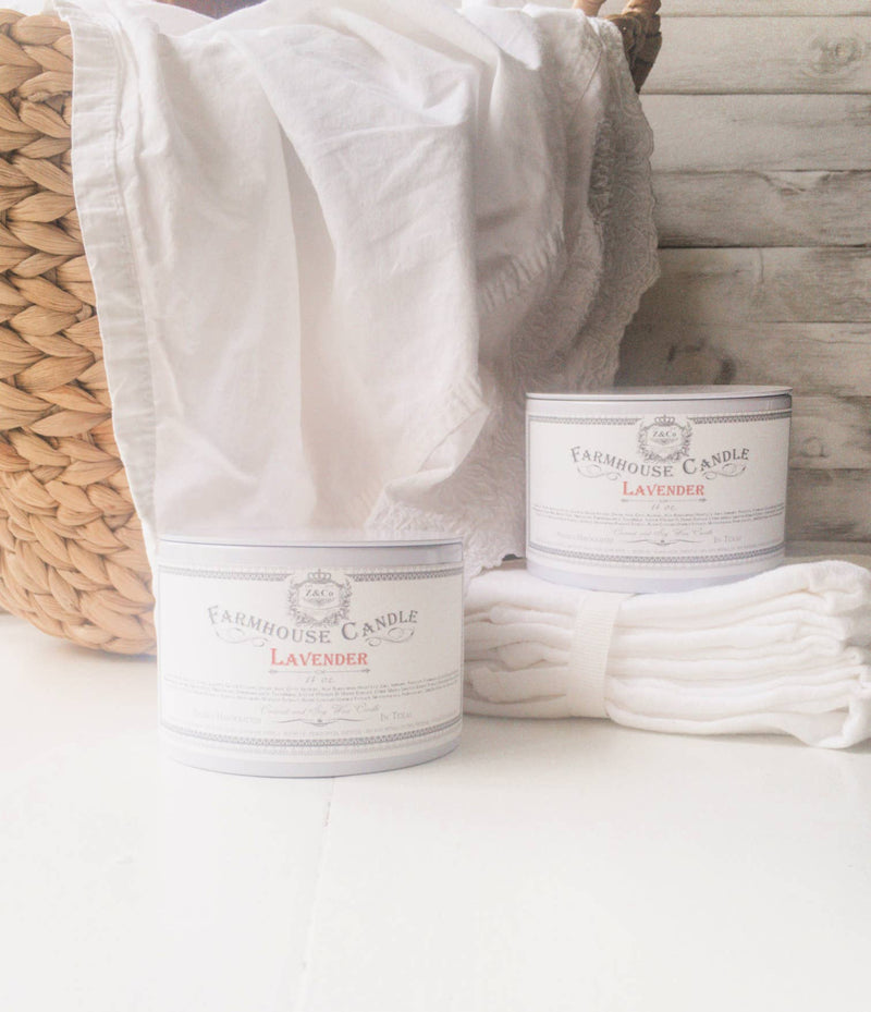 Two Z&Co. Lavender Farmhouse Tin Candles made with natural vegetable wax, next to a white cloth and a wicker basket, set against a white wooden backdrop.