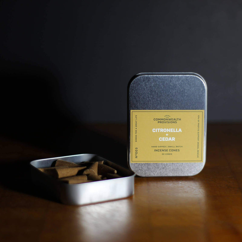 A tin container of Commonwealth Provisions Citronella + Cedar Incense Cones on a wooden table, with the lid open showing the contents, and a label visible in a dimly lit room, ideal for outdoor use.