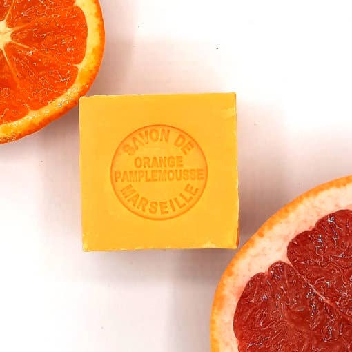 A vibrant orange Senteurs De France Marseille Orange Grapefruit Cube soap with embossed text "Savon de orange pamplemousse" surrounded by sliced halves of an orange and a grapefruit on a white background, certified by the French Soap.