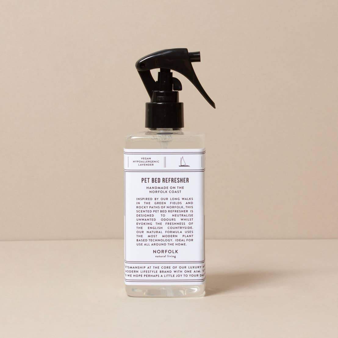 A clear plastic spray bottle featuring a black nozzle is set against a beige backdrop. The label, "Norfolk Natural Living Lavender Pet Bed Refresher," emphasizes its antibacterial and odor-fighting capabilities, along with information about its usage and ingredients.