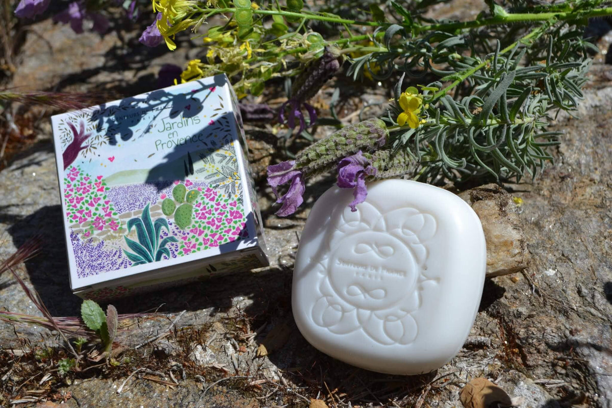 A bar of Senteurs De France Lavender Soap "Gardens of Provence" beside a small, illustrated book titled "jardins de provence", placed on a rocky surface with natural vegetation around.