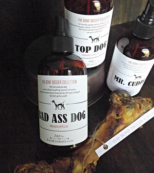 Three bottles labeled "top dog," "Z&Co. Bad Ass Dog Spray Mist," and "mr. cuddles" from the bone digger collection, placed next to a chewed bone on a wooden surface, all featuring