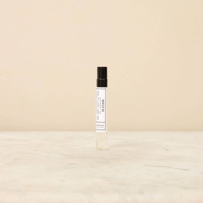 A small, clear glass rollerball bottle of Norfolk Natural Living - Days of Winter 90 Parfum 10ml, possibly floral notes perfume or essential oil, labeled xoxoux, stands against a neutral light beige background.