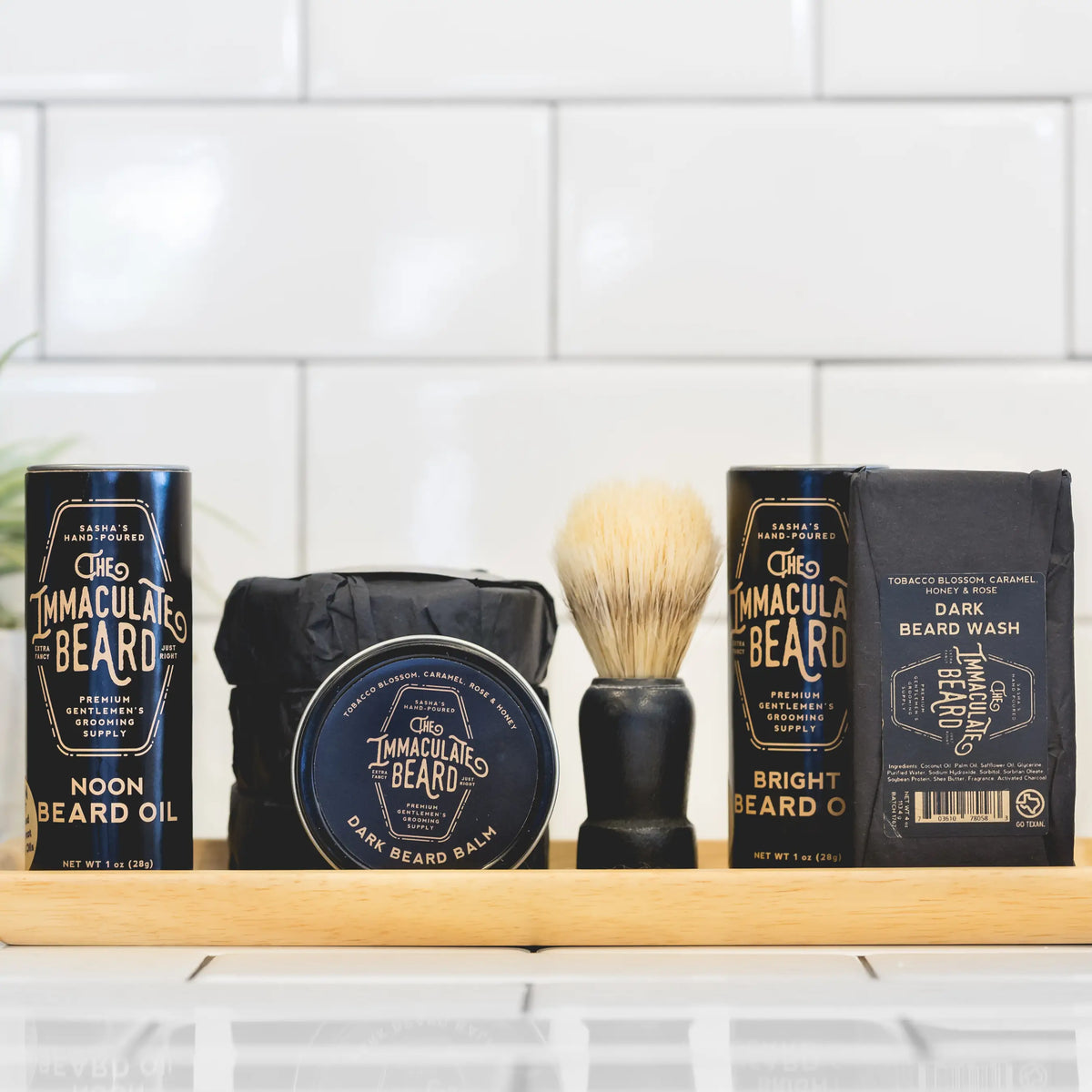 A range of The Immaculate Beard men's grooming products on a wooden shelf against a white tiled background, including beard oil, beard wash, shaving brush, The Immaculate Beard - Shave Soap Puck, and other packaged products.