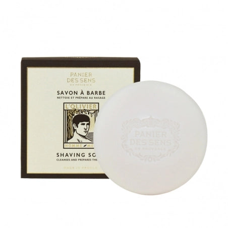 A round, white Panier des Sens L’Olivier Shaving Soap next to its packaging, which features vintage-style graphics and text that includes the brand name "panier des sens" and product details.