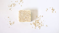 A bar of Homegrown {77833} Co - Bi*** Better Have My Honey soap surrounded by loose oat flakes on a white background.