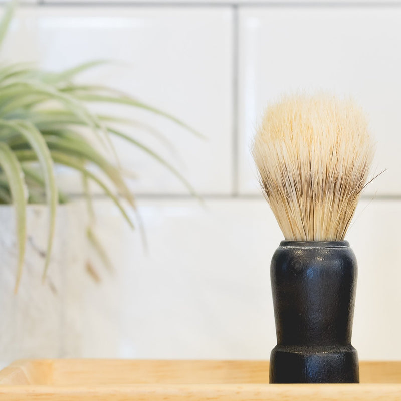 The Immaculate Beard - Shave Brush with a black handle on a wooden surface, with a green potted plant and white tiled wall in the background.