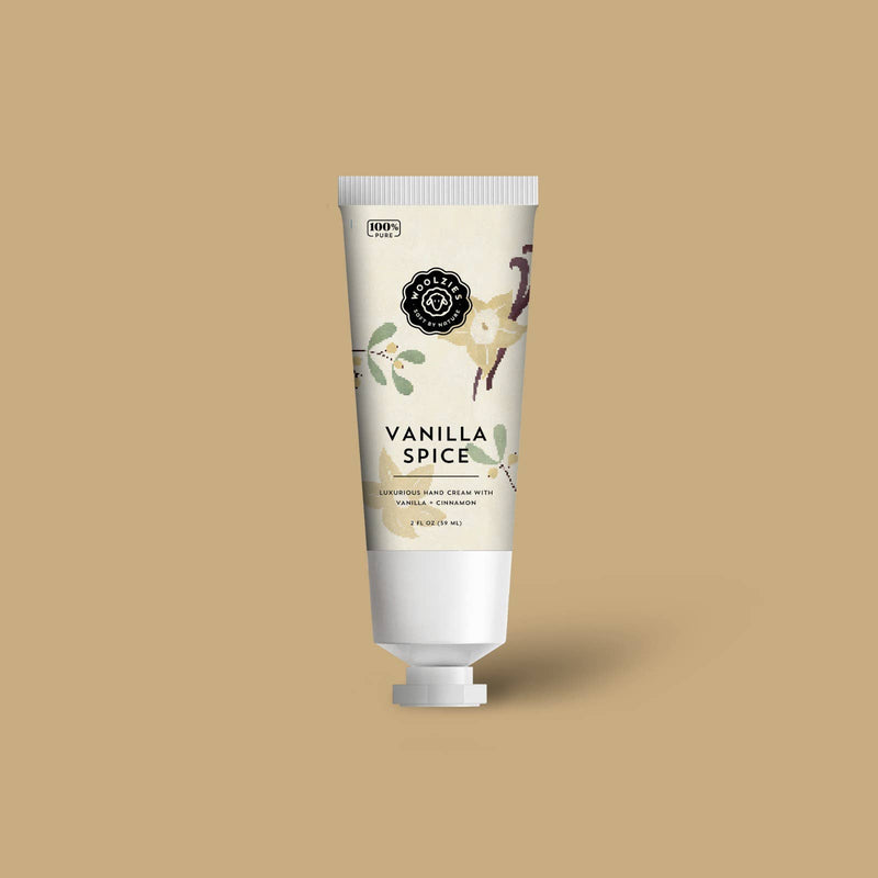 A tube of Woolzies Vanilla Spice Hand Cream 2oz with shea butter on a plain beige background. The tube features floral and vanilla pod illustrations with elegant black and white labeling.