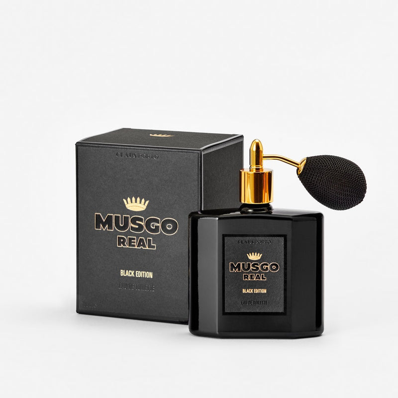 A black, square bottle of Claus Porto Musgo Real Black Edition EDT with a gold spray bulb, accompanied by its matching box, against a white background.