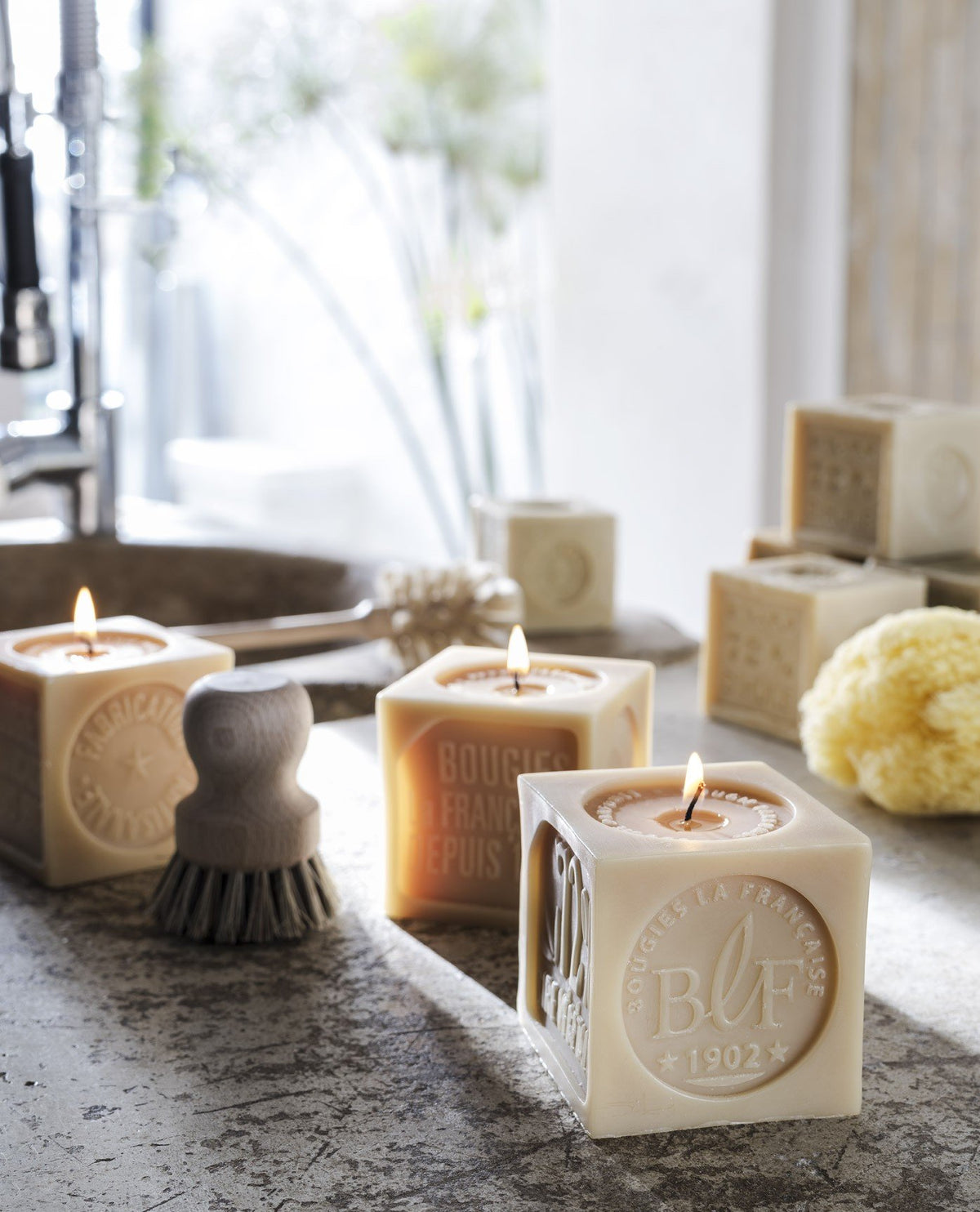 Three lit square candles with embossed lettering on a bathroom vanity, complemented by a sponge and toiletries, including a Bougies la Française Marseille Soap Decorative Candle, in a serene setting with natural light.