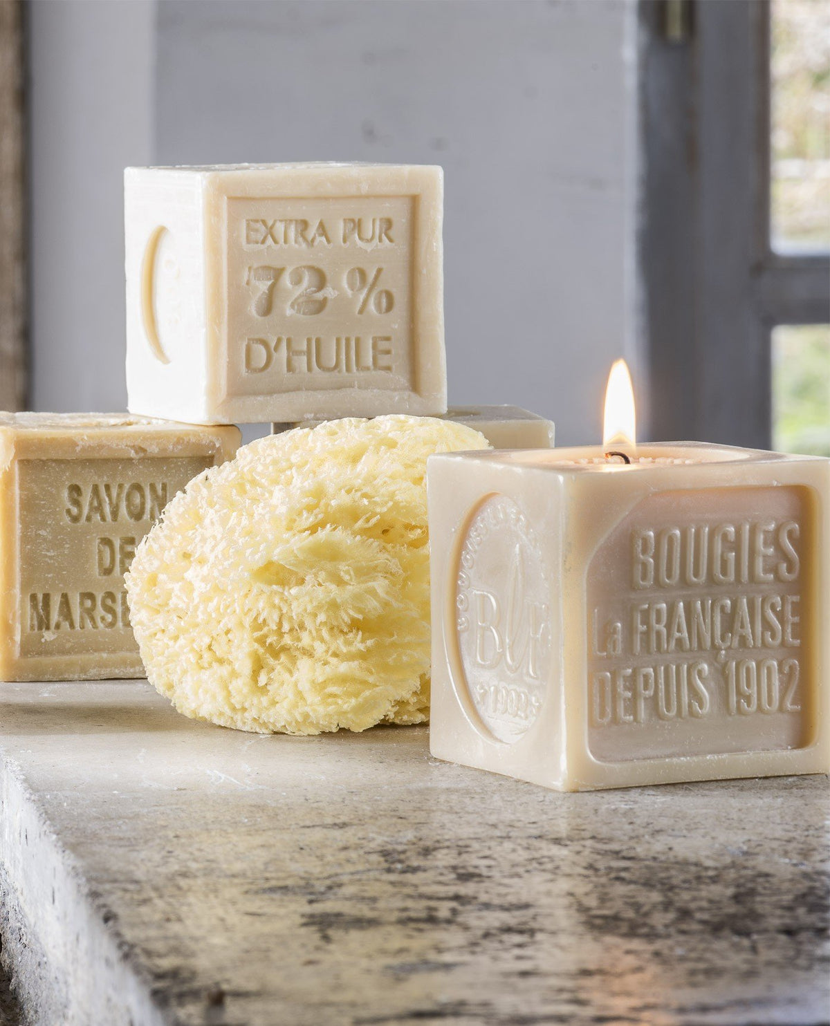 Lit Bougies la Française decorative candle next to bars of Marseille soap and a natural sponge on a stone windowsill, with a garden view in the background.