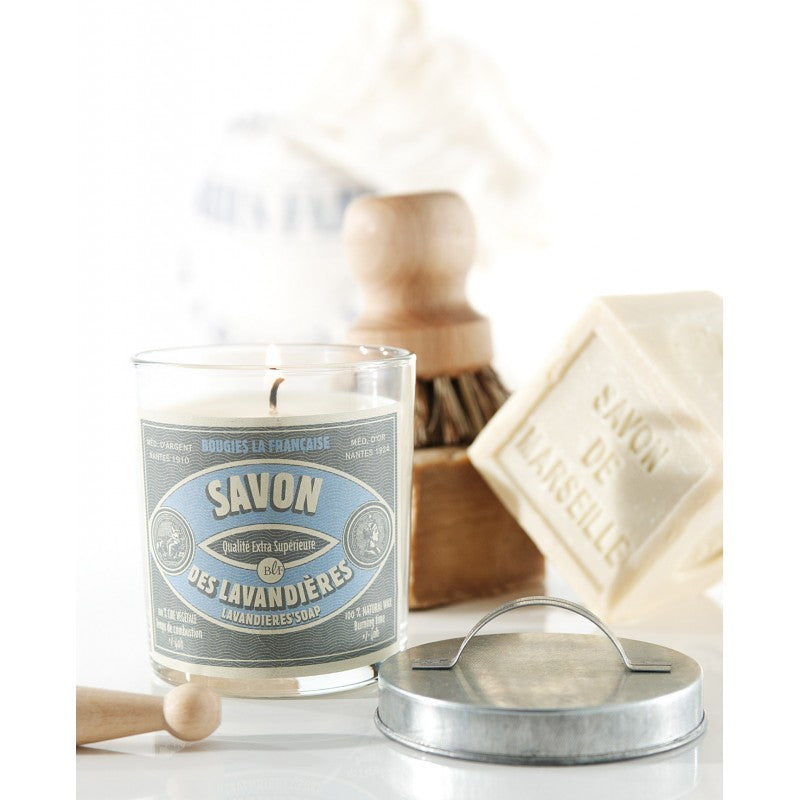 A Bougies la Francaise Artisan Lavandières Soap Candle with a vintage label that reads "savon des lavandières," next to a bar of lavender soap with the same label, a scrub brush, and a tin container, set
