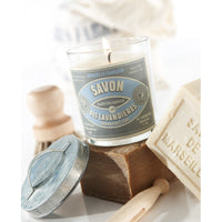 A Bougies la Francaise Artisan Lavandières soap candle in a clear glass holder labeled "savon des lavandieres" beside traditional soap bars and a wooden brush, all on a bright, soft background.
