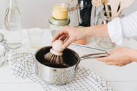 A person using an Andrée Jardin Tradition Saucepan Brush made in France by Andrée Jardin to stir in a saucepan on a kitchen counter, surrounded by various kitchen bottles and containers.