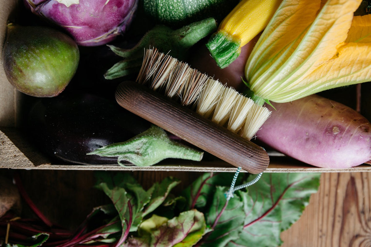 A variety of fresh vegetables including zucchini, eggplants, and leafy greens in a wooden box with an Andrée Jardin Hard & Soft Vegetable Brush Heritage made of natural fibers resting on top.